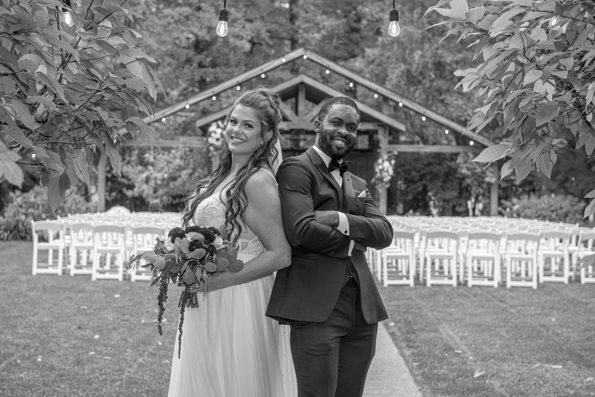 A smiling bride and groom stand back-to-back outdoors, surrounded by white chairs and string lights under leafy branches.