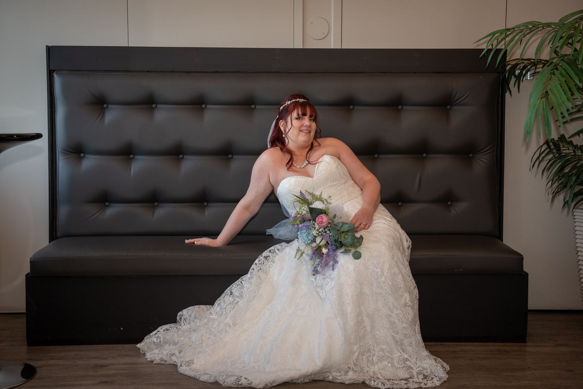 A bride in a white lace gown sits playfully on a dark tufted bench, holding a bouquet of purple and green flowers.