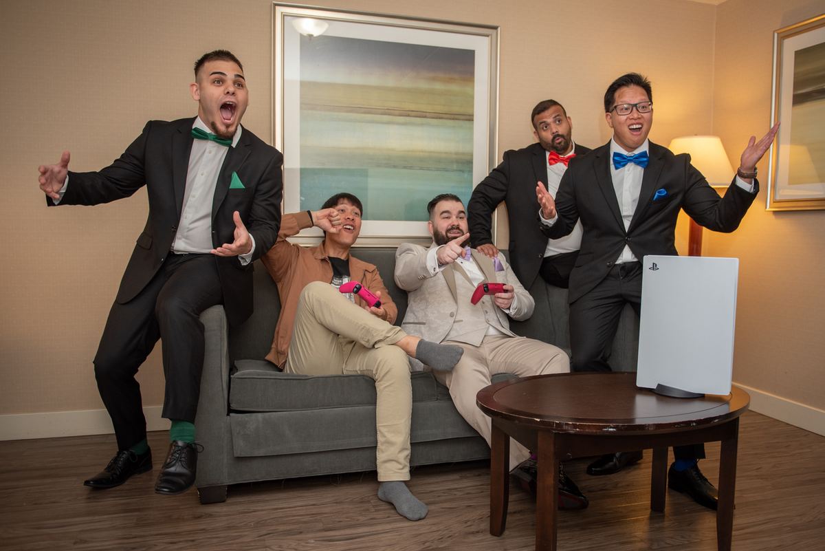 Five men in suits and bow ties celebrate an exciting moment while playing video games, with one jumping from the couch.