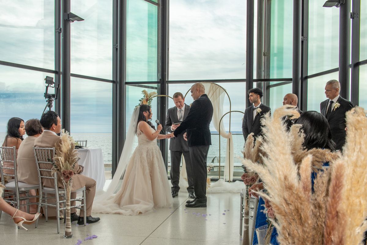 A bride and groom stand at an altar with large windows behind them framing the ocean, guests seated in rows.