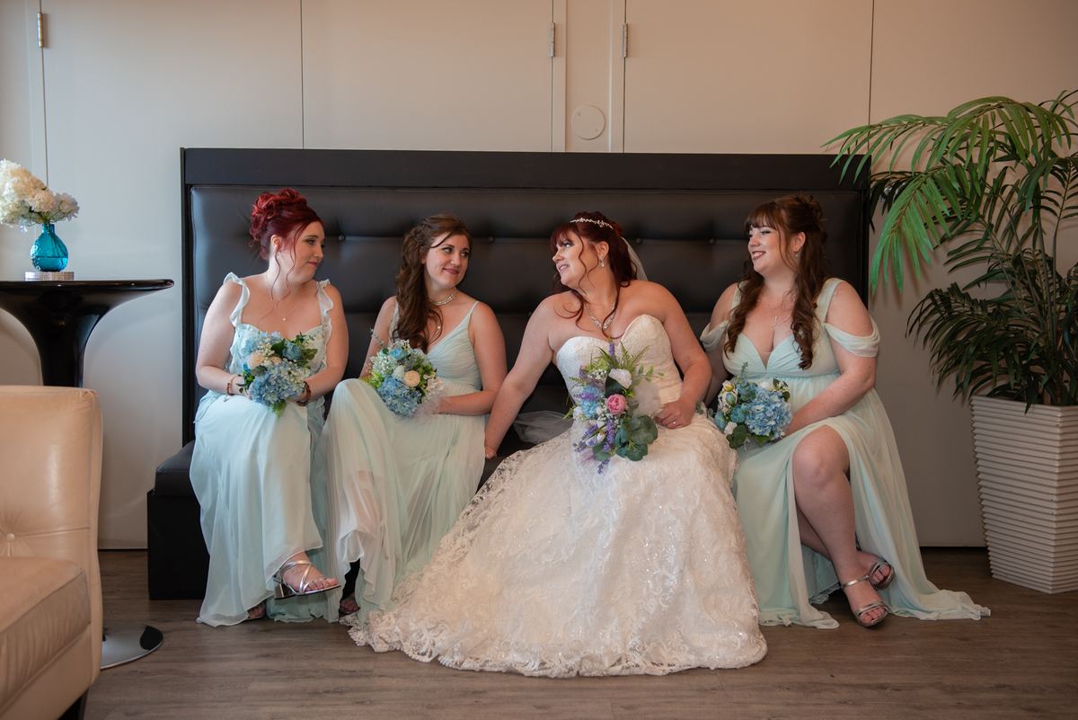 Four women, including the bride in a white lace gown, sit on a dark headboard laughing and looking at each other.