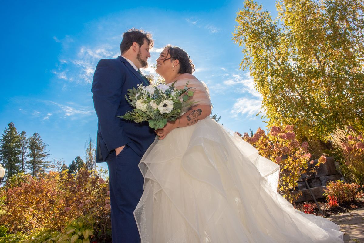 A bride in white and groom in black laugh together outdoors under blue sky, surrounded by autumn leaves and flowers.