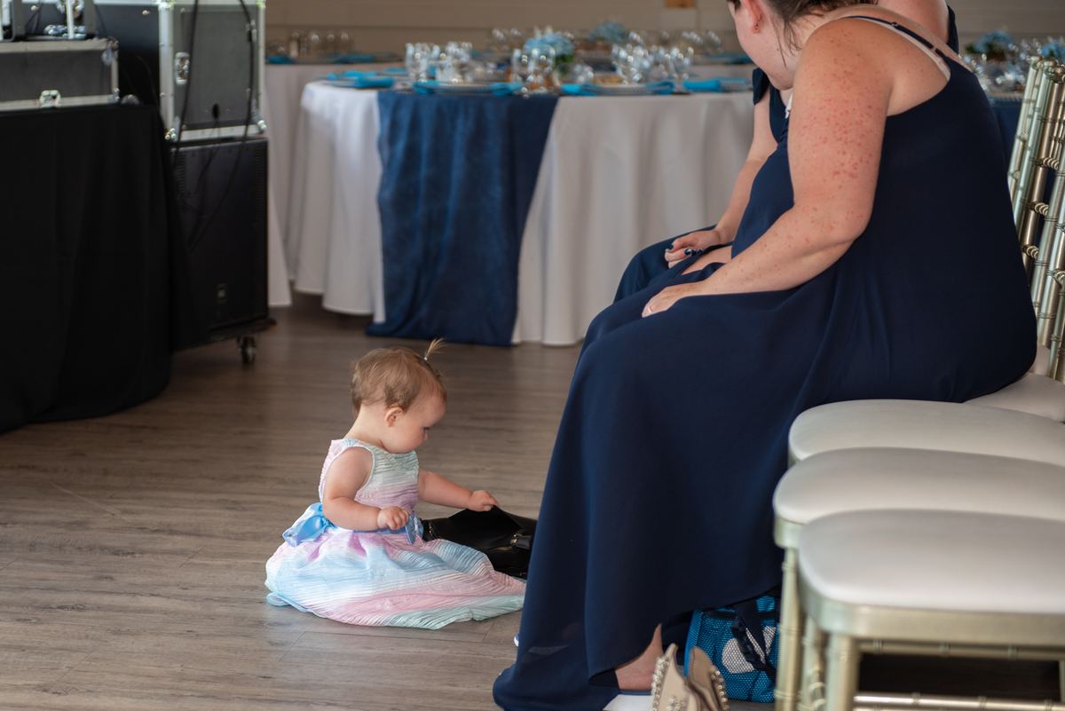 A young child in a pastel gown sits on the floor, exploring a black bag at the feet of a woman seated in a navy dress.