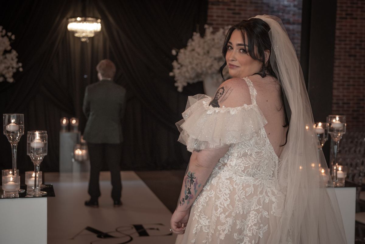 A bride in an off-the-shoulder lace gown smiles over her shoulder at the altar, softly lit by candles and a chandelier.