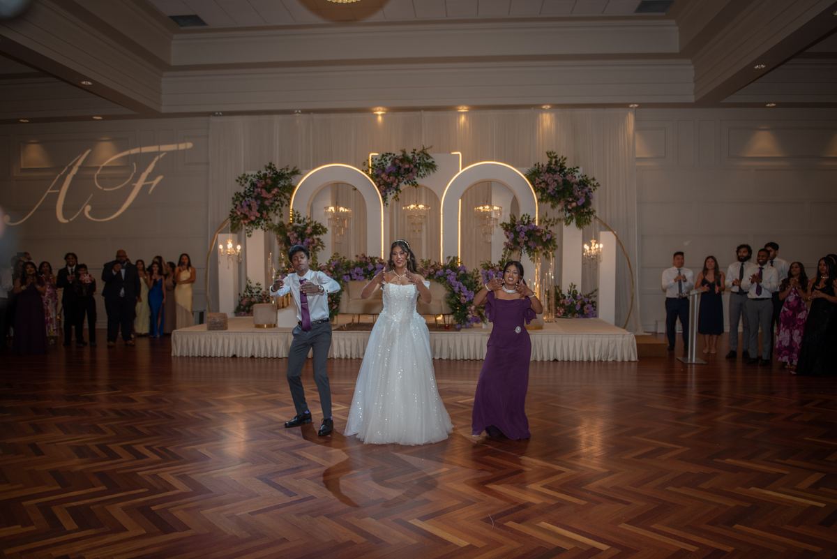 A bride in a sparkling white gown dances with her groom on a hardwood floor as guests clap beneath flower-covered arches.