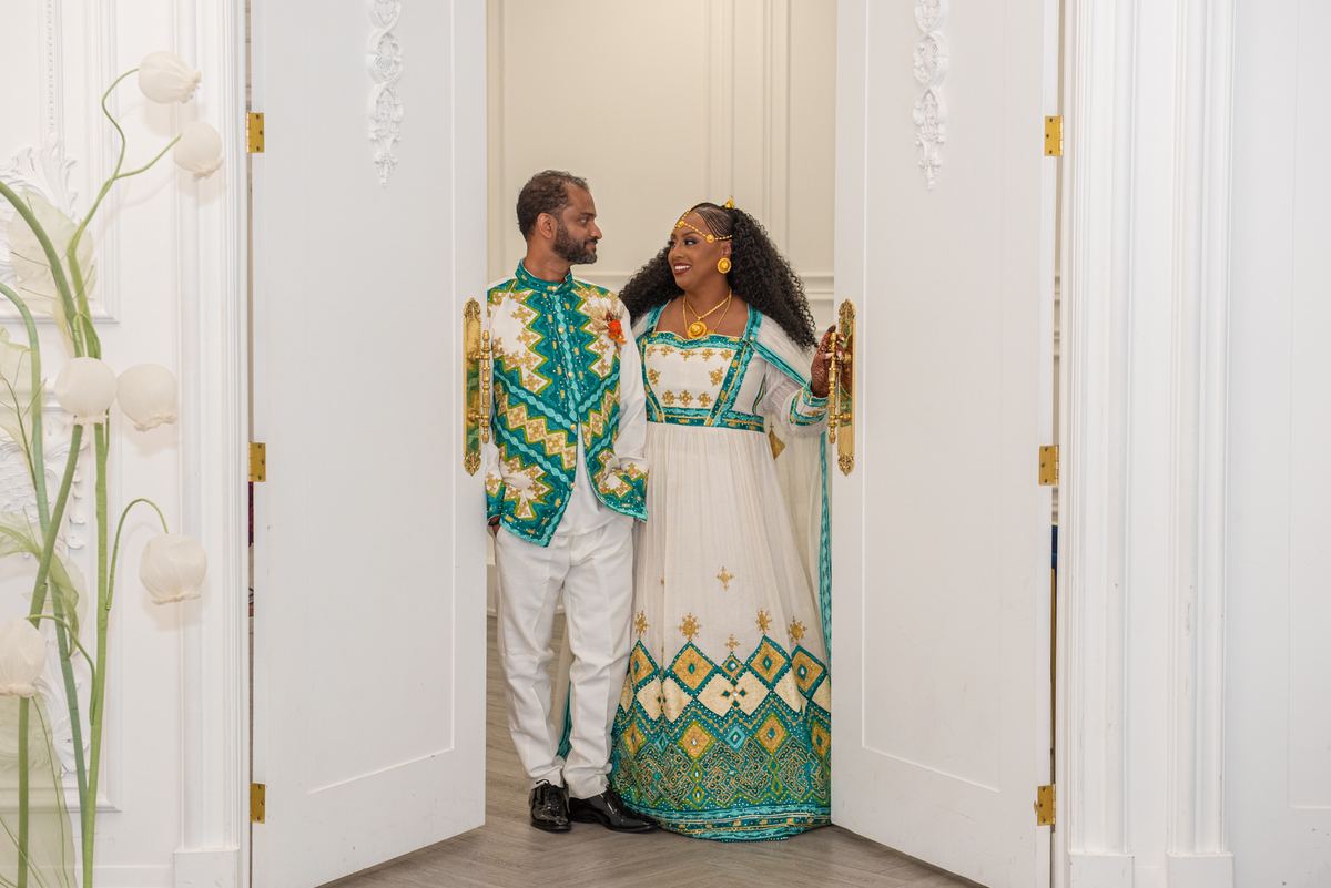 A joyful couple in ornate traditional attire stands by an open white doorway, gazing lovingly at each other.