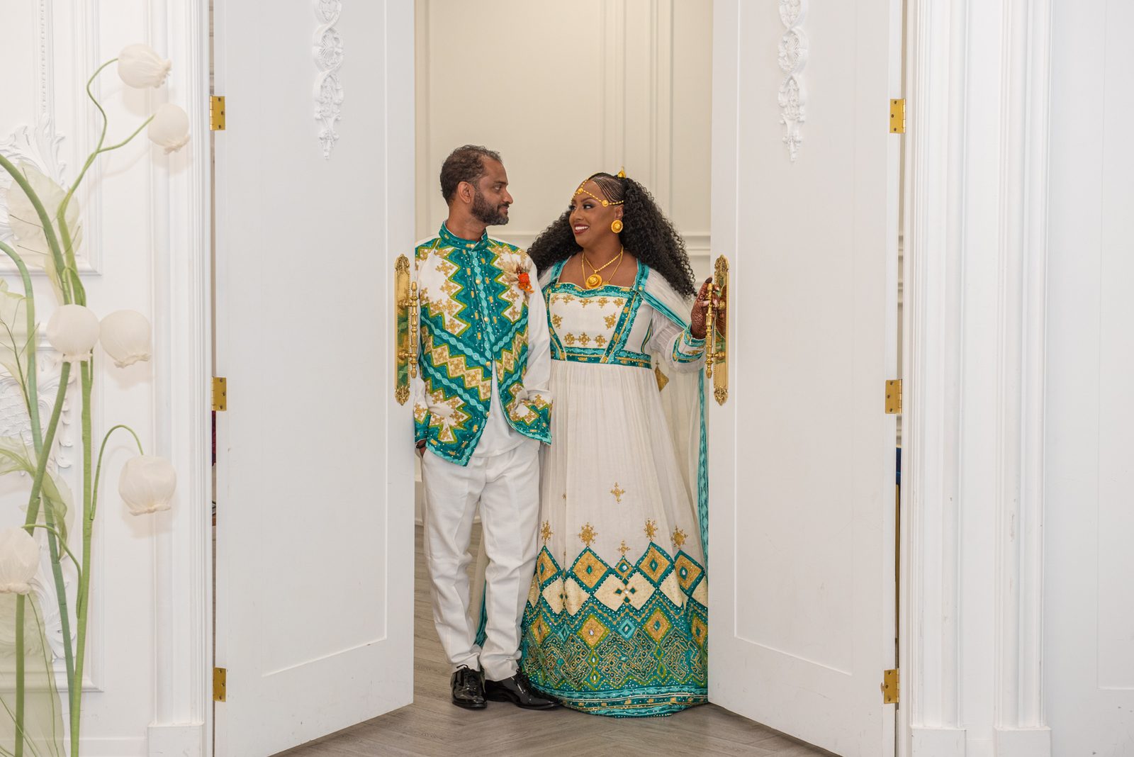 A joyful couple in ornate traditional attire stands by an open white doorway, gazing lovingly at each other.