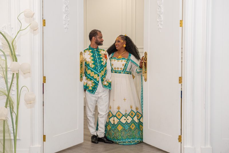 A joyful couple in ornate traditional attire stands by an open white doorway, gazing lovingly at each other.