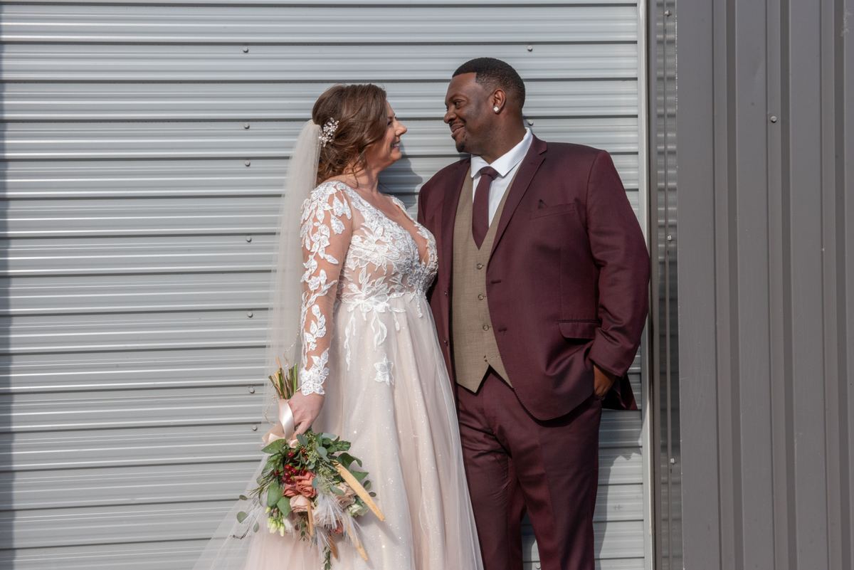 A bride and groom smile warmly at each other against a metal backdrop on their wedding day, illuminated by sunlight.
