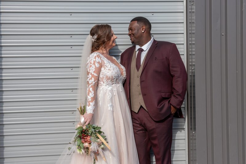 A bride and groom smile warmly at each other against a metal backdrop on their wedding day, illuminated by sunlight.