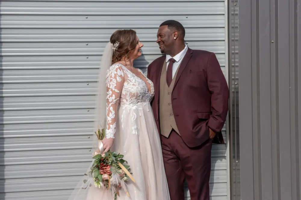 A bride and groom smile warmly at each other against a metal backdrop on their wedding day, illuminated by sunlight.
