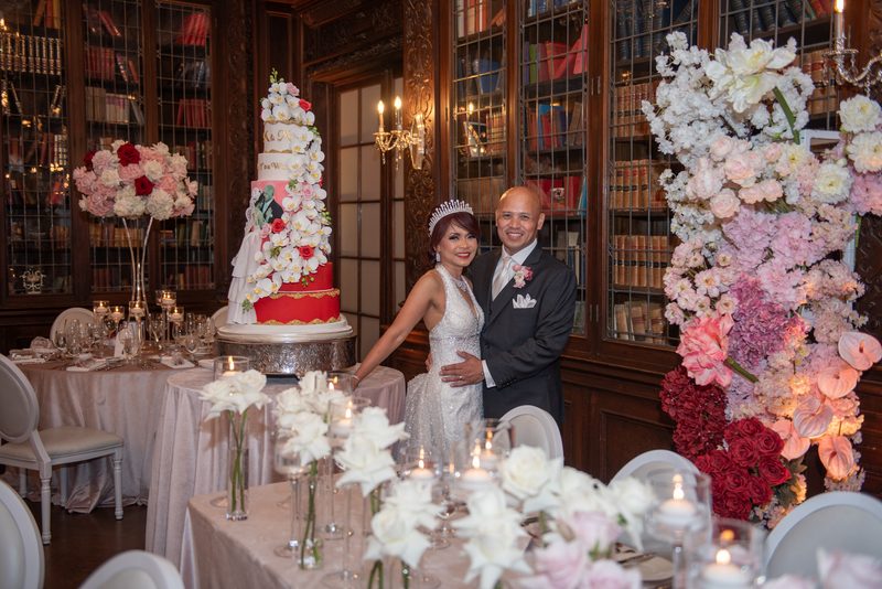 A newlywed couple smiles in a lavish library setting with floral arrangements and a grand wedding cake.