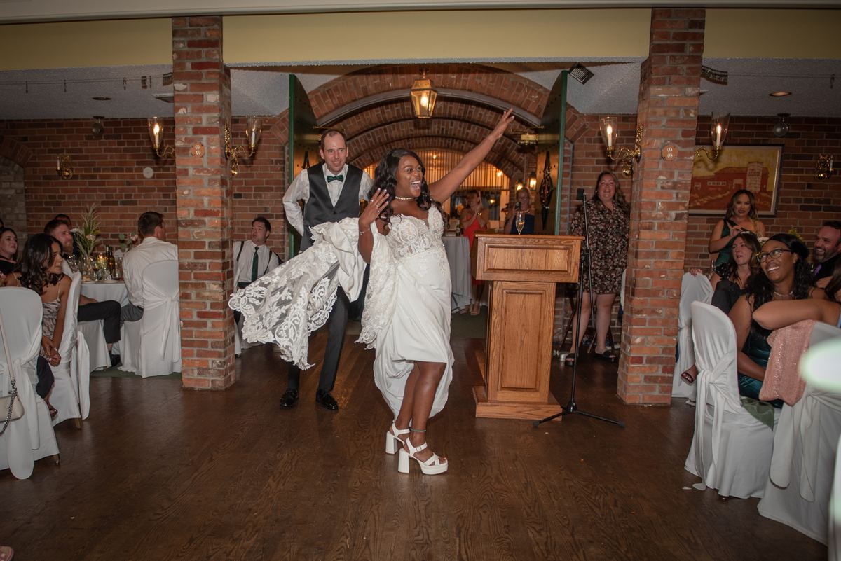 A bride in a white gown dances with her arms raised as her groom holds up her train, surrounded by smiling guests.