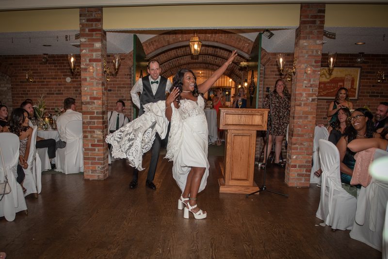 A bride in a white gown dances with her arms raised as her groom holds up her train, surrounded by smiling guests.
