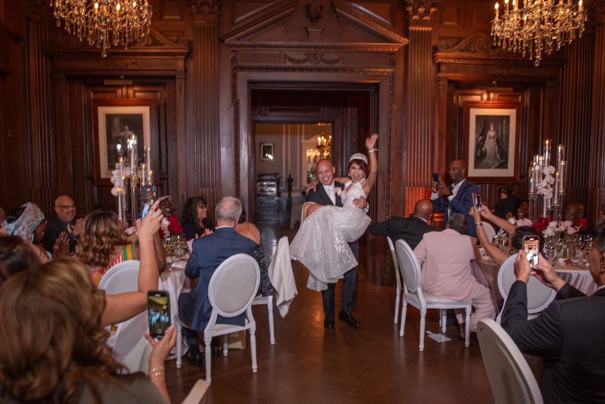 A groom lifts his bride in a wood-paneled ballroom, surrounded by guests on phones, festive atmosphere.