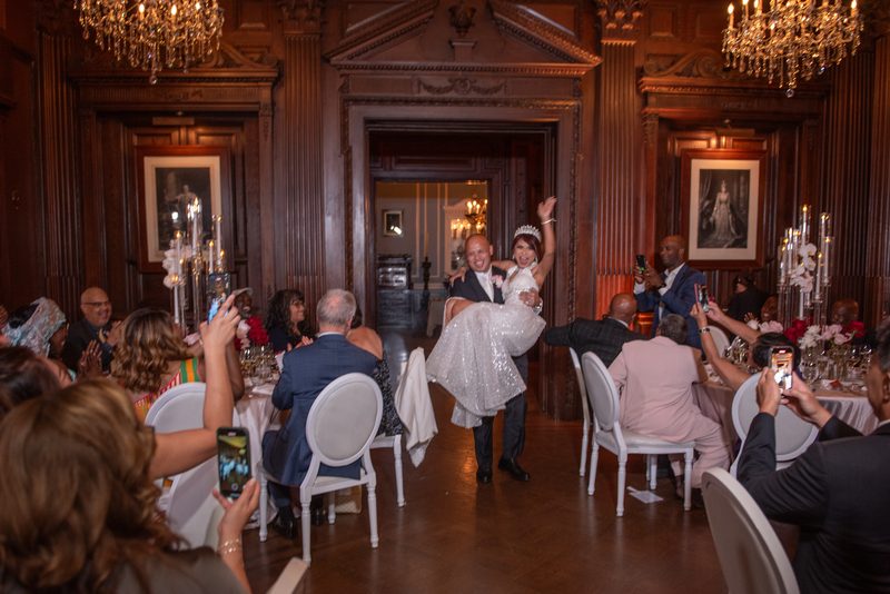 A groom lifts his bride in a wood-paneled ballroom, surrounded by guests on phones, festive atmosphere.