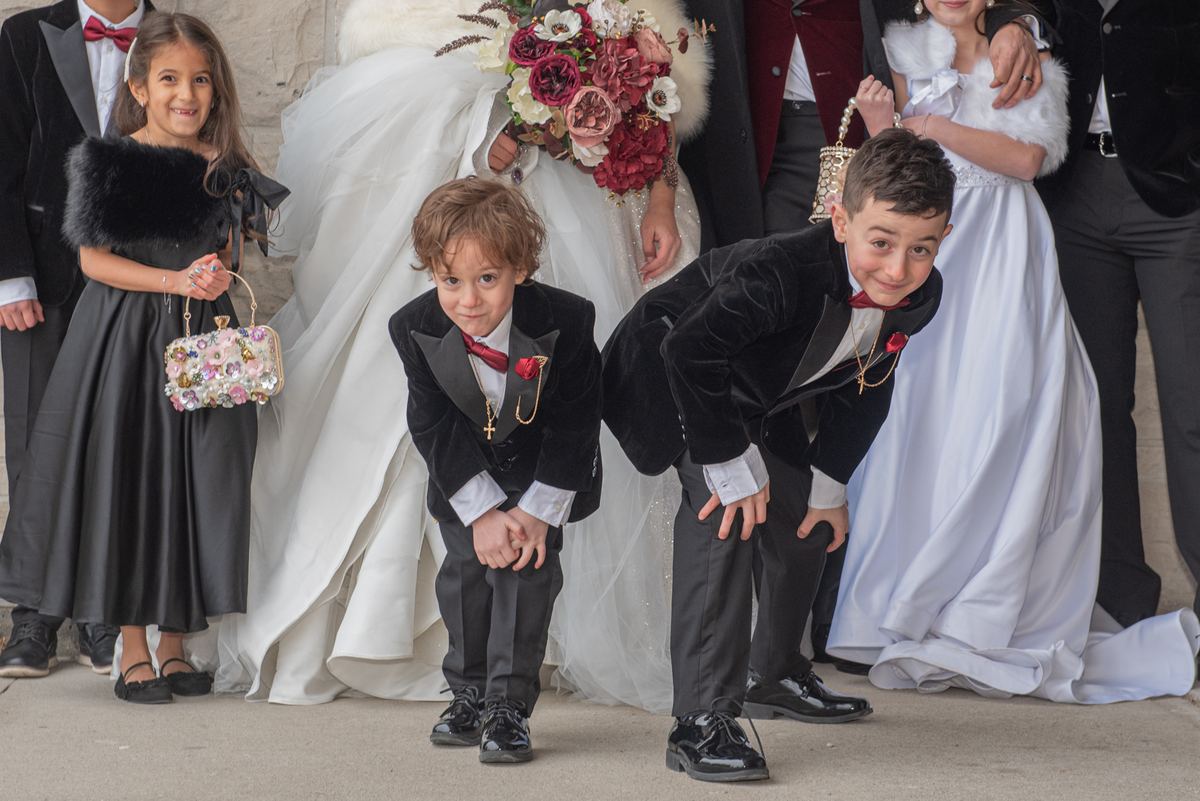 Two young boys in black velvet suits playfully crouching, with a girl and bride behind them at a wedding.