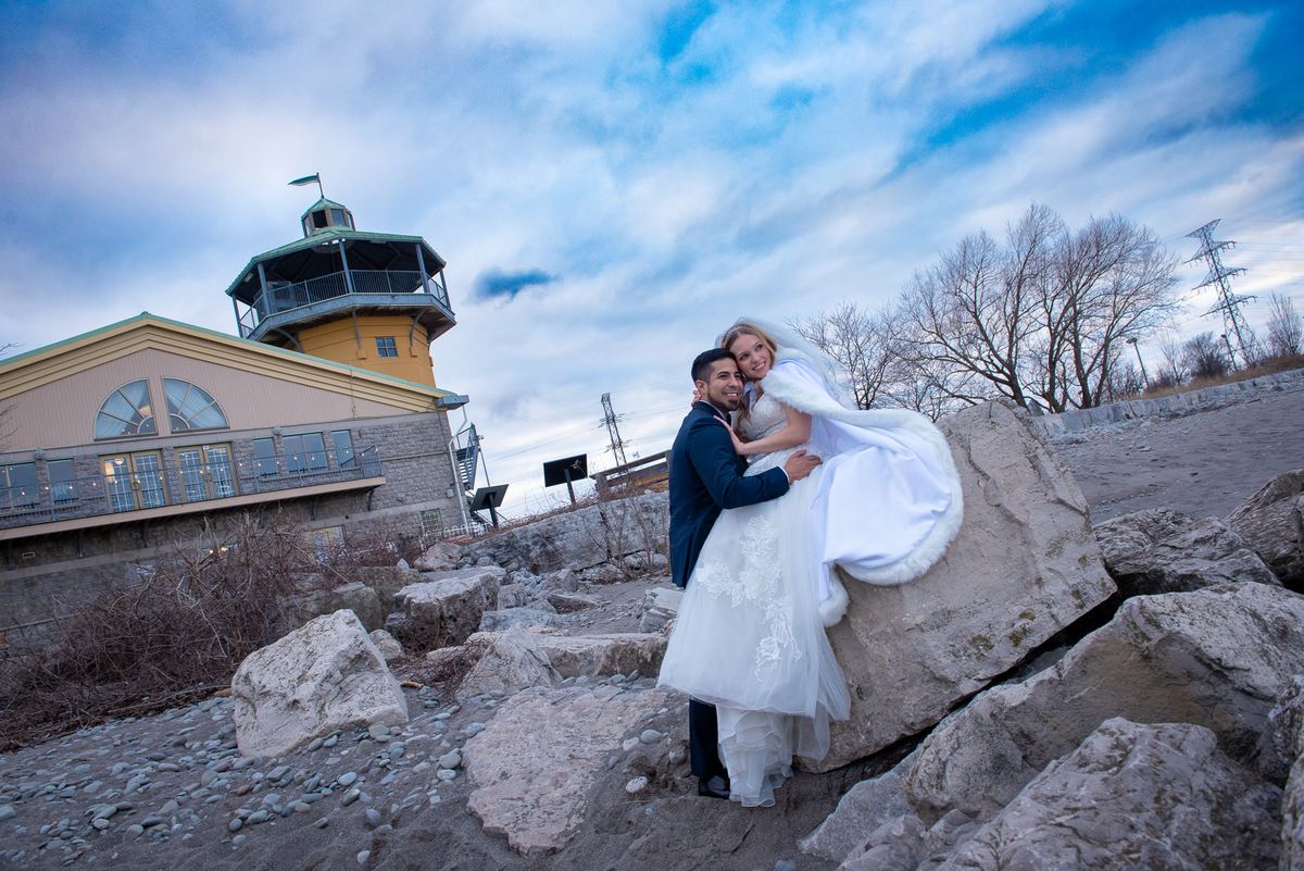 A bride and groom embrace on a rocky waterfront with a lighthouse in the background under a partly cloudy sky.