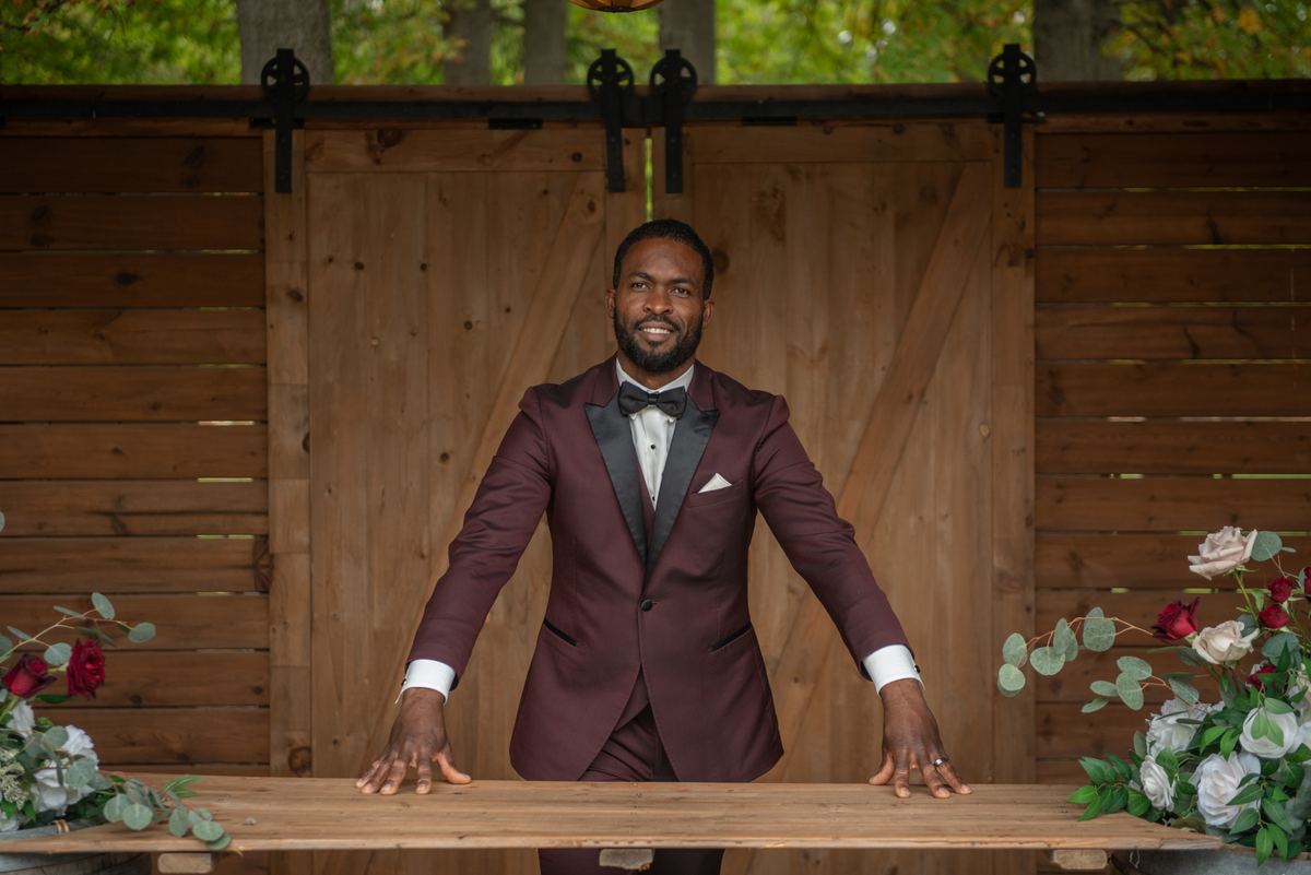 A smiling Black man in a burgundy tuxedo stands behind a wooden table with floral arrangements, under trees.