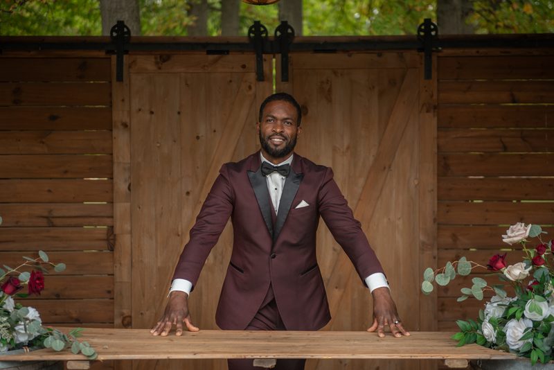 A smiling Black man in a burgundy tuxedo stands behind a wooden table with floral arrangements, under trees.