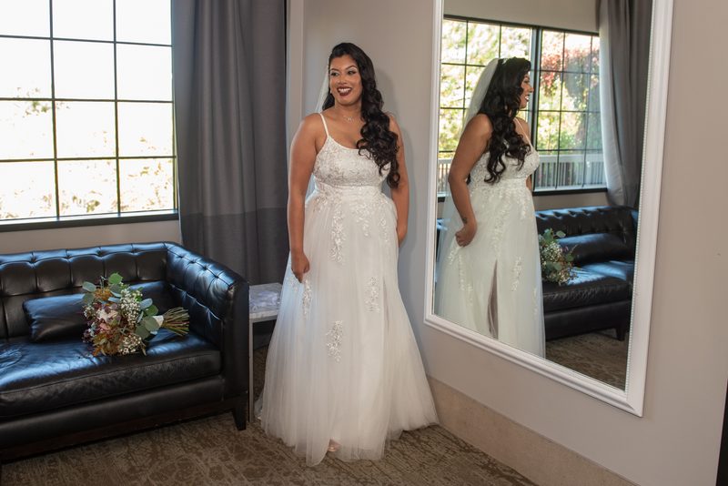 A bride smiles at her reflection in a mirror, wearing an elegant white gown with lace detailing near a bouquet.