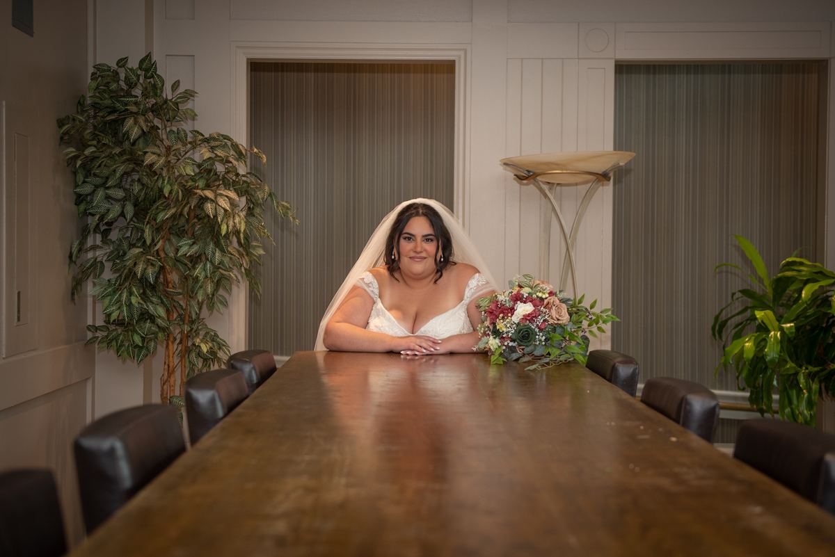 A smiling bride rests her arms on a wooden table with her bouquet, in a softly lit room with potted plants and modern chairs.