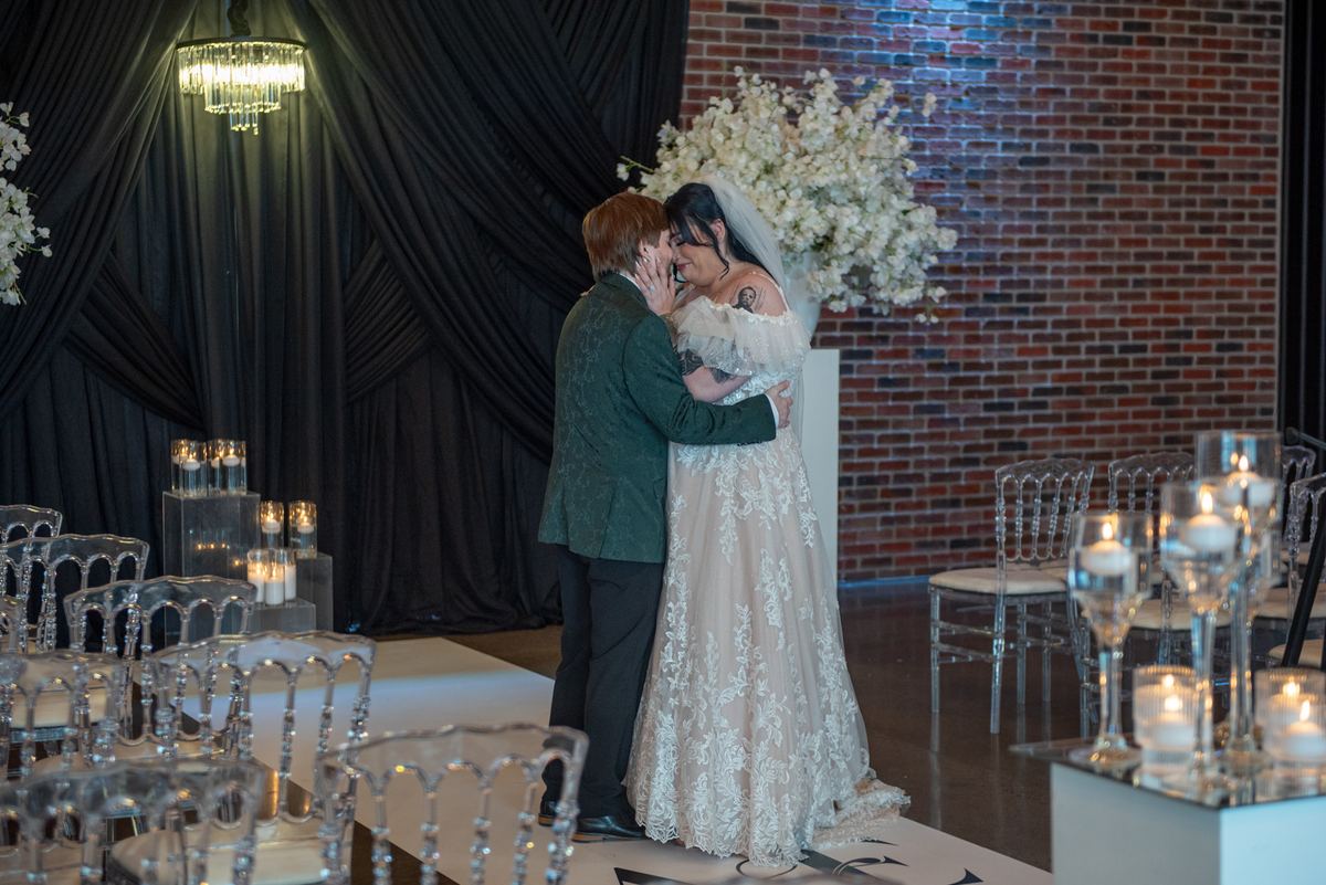 A bride and groom embrace intimately by candlelight, their faces close against a backdrop of black drapes and exposed brick.