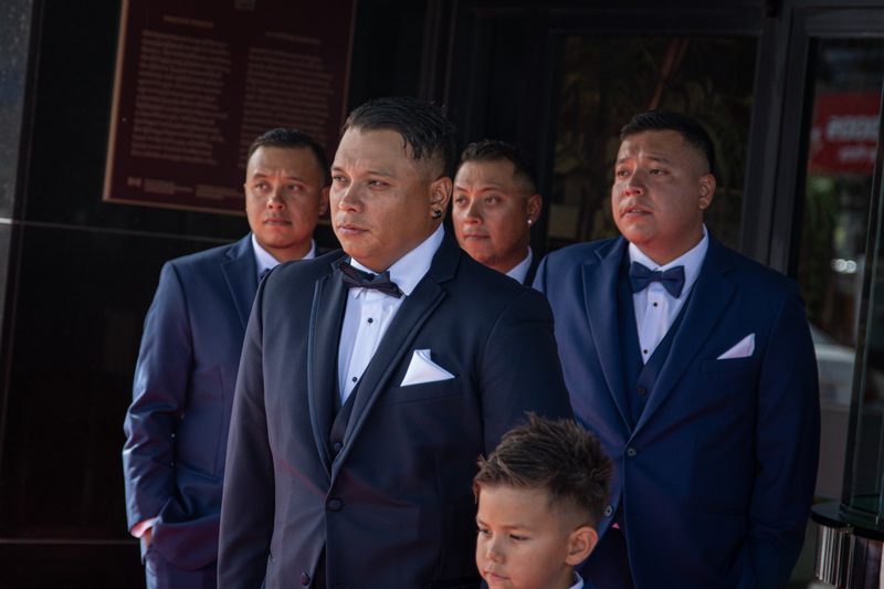 Men in navy tuxedos stand solemnly outdoors as a young boy is visible in the foreground during daylight.