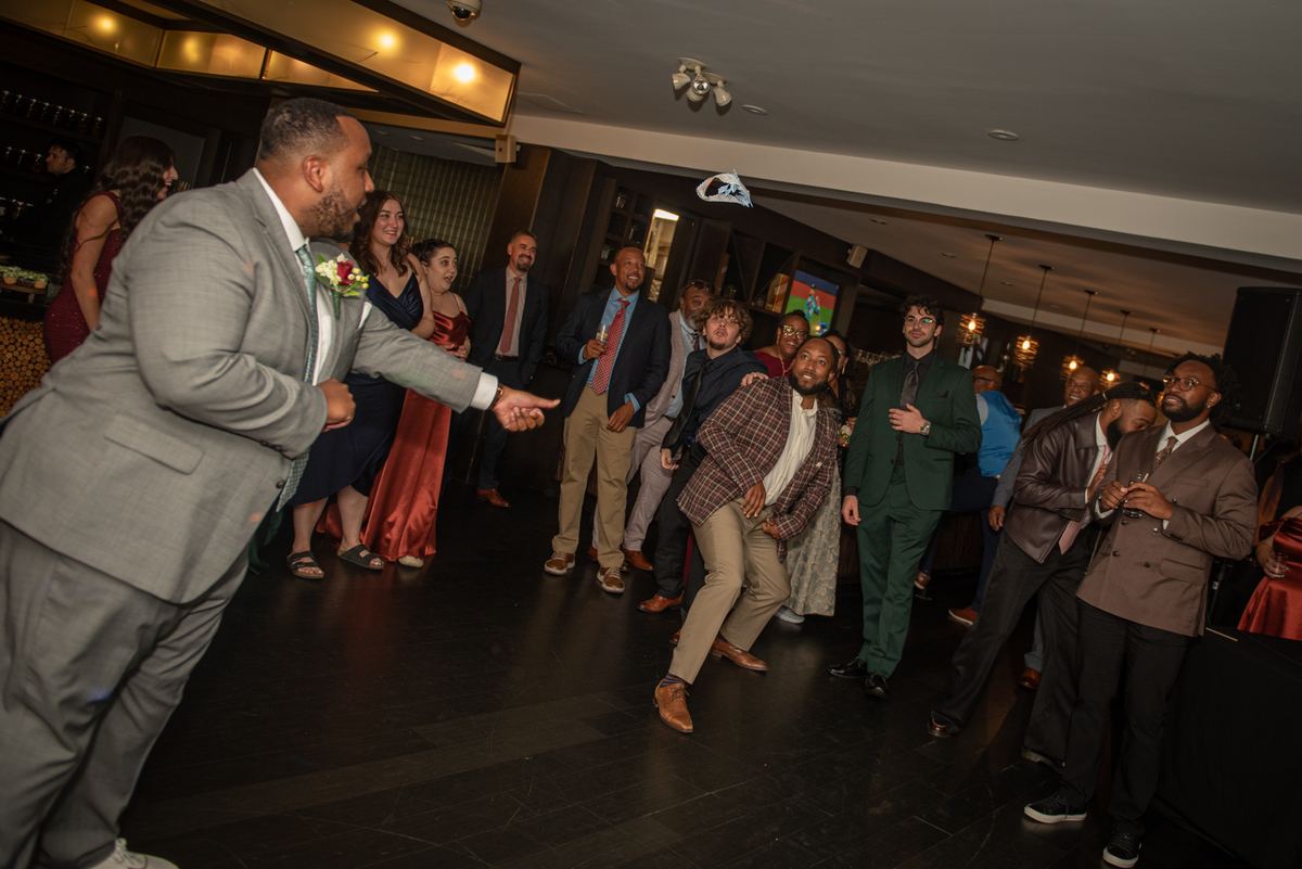 A groom in a gray suit tosses his hat into the air, eliciting laughter from formally dressed guests at a wedding reception.