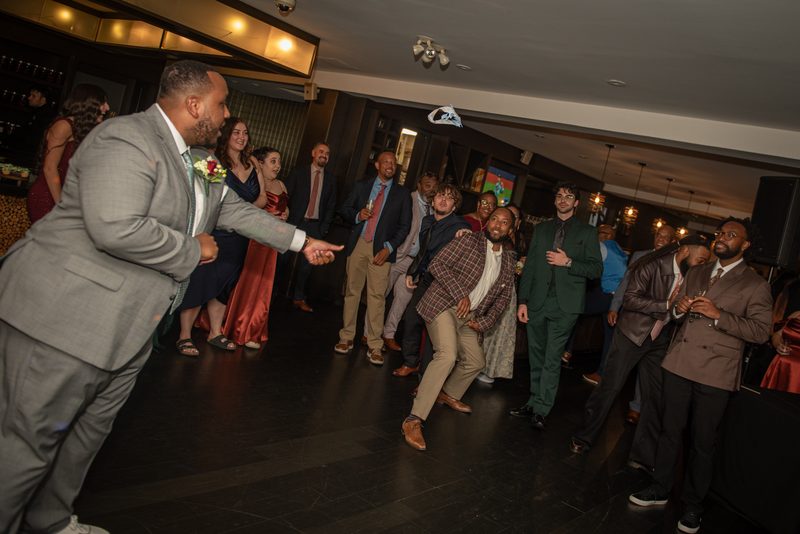 A groom in a gray suit tosses his hat into the air, eliciting laughter from formally dressed guests at a wedding reception.