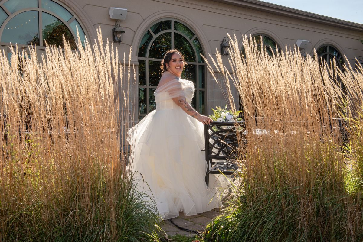 A smiling bride in a white gown stands among golden grasses, holding flowers near an arched windowed building.