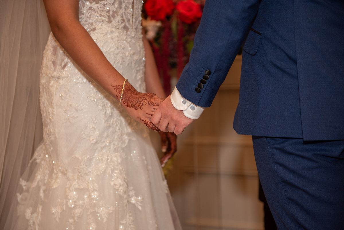 Bride and groom hold hands, her henna design visible on her hand, his navy suit formal against the lace of her gown.