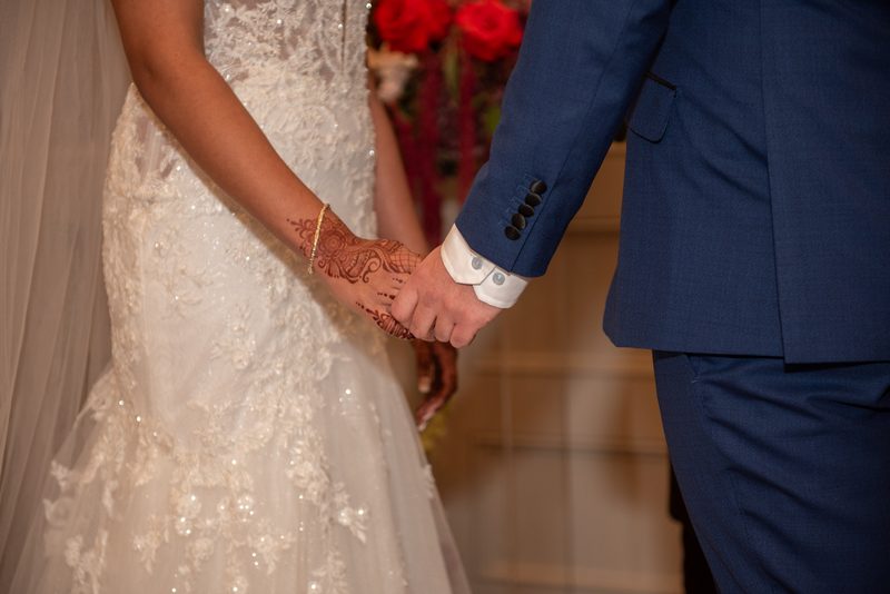 Bride and groom hold hands, her henna design visible on her hand, his navy suit formal against the lace of her gown.