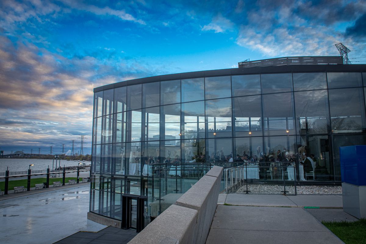 A modern glass-walled event space by the waterfront during twilight with guests seated inside at tables.
