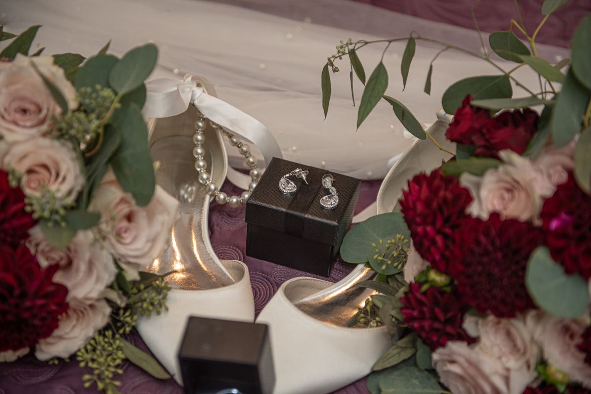 White high-heeled shoes, sparkling earrings in a black box, and floral arrangements of roses and eucalyptus.