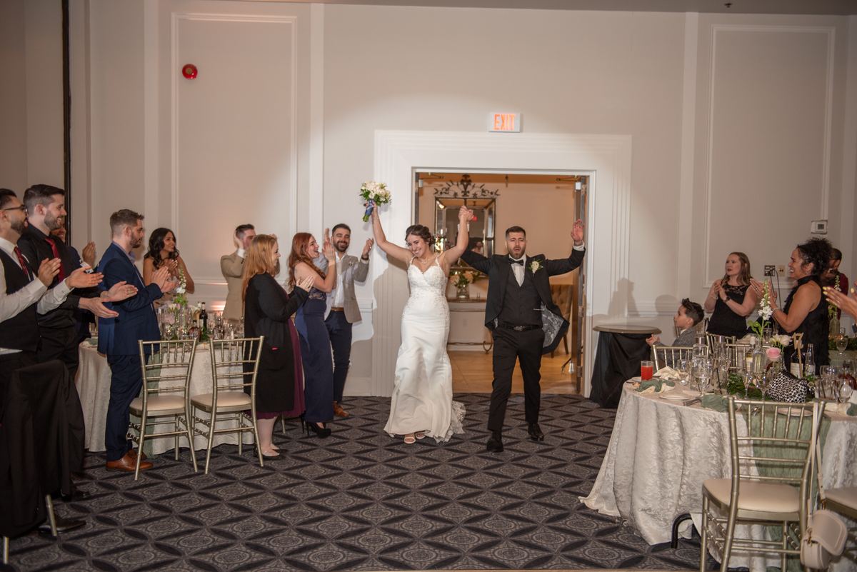 Bride and groom walk down the aisle in a banquet hall, arms raised as guests applaud, spotlight illuminating them.