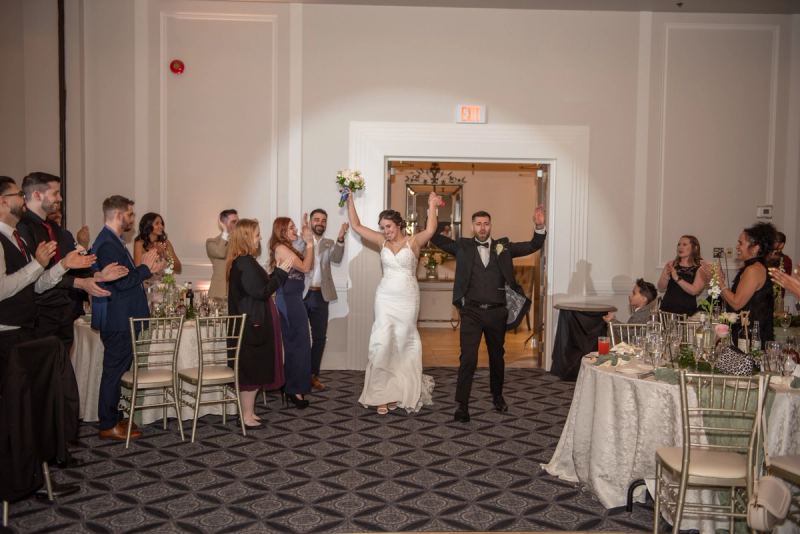 Bride and groom walk down the aisle in a banquet hall, arms raised as guests applaud, spotlight illuminating them.