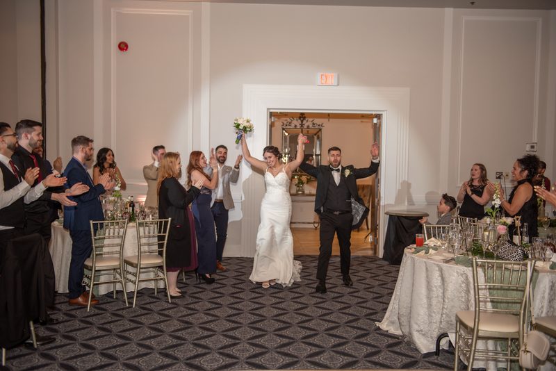 Bride and groom walk down the aisle in a banquet hall, arms raised as guests applaud, spotlight illuminating them.