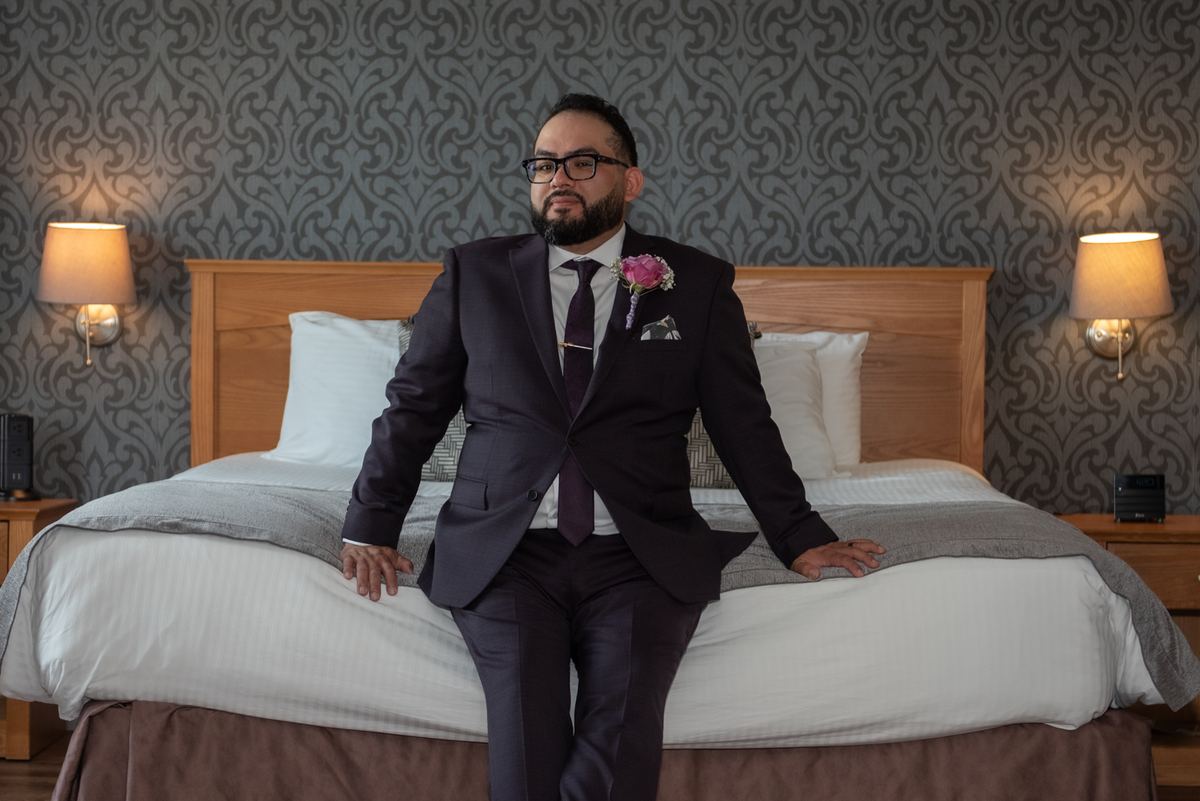 A groom in a dark suit sits confidently on a hotel bed, hands resting on the mattress, against patterned gray wallpaper.