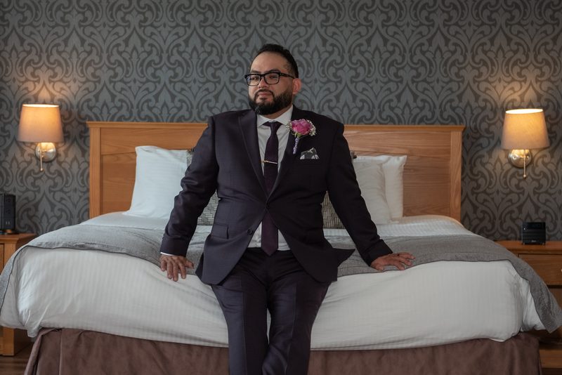 A groom in a dark suit sits confidently on a hotel bed, hands resting on the mattress, against patterned gray wallpaper.
