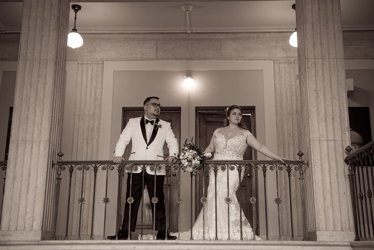 A bride in a lace gown and groom in a tuxedo stand on an ornate balcony framed by classical columns under warm lighting.