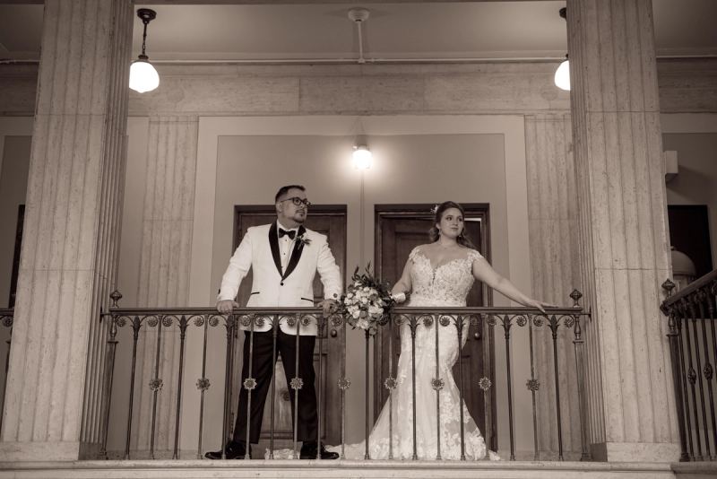 A bride in a lace gown and groom in a tuxedo stand on an ornate balcony framed by classical columns under warm lighting.