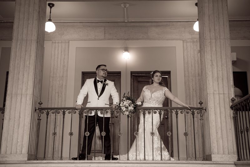 A bride in a lace gown and groom in a tuxedo stand on an ornate balcony framed by classical columns under warm lighting.