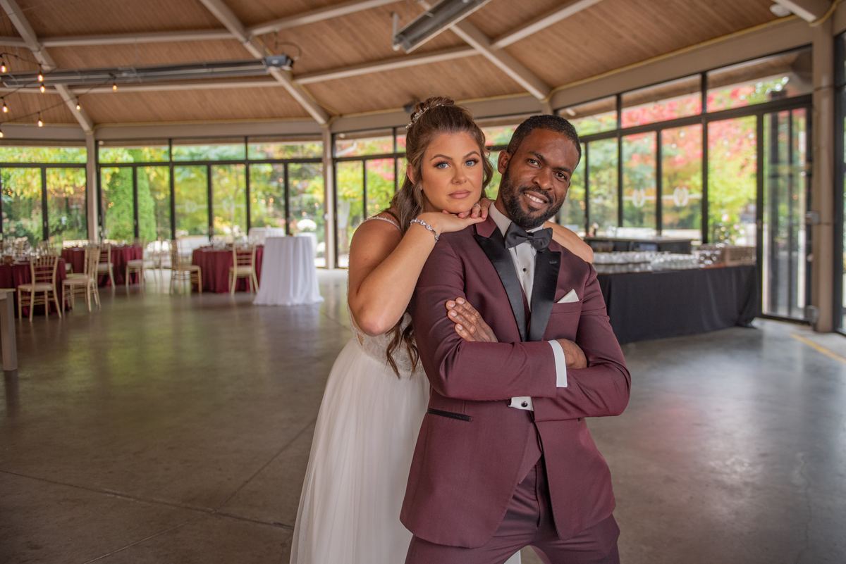 A bride and groom pose intimately in a modern hall with autumn foliage visible through large windows, under soft light.