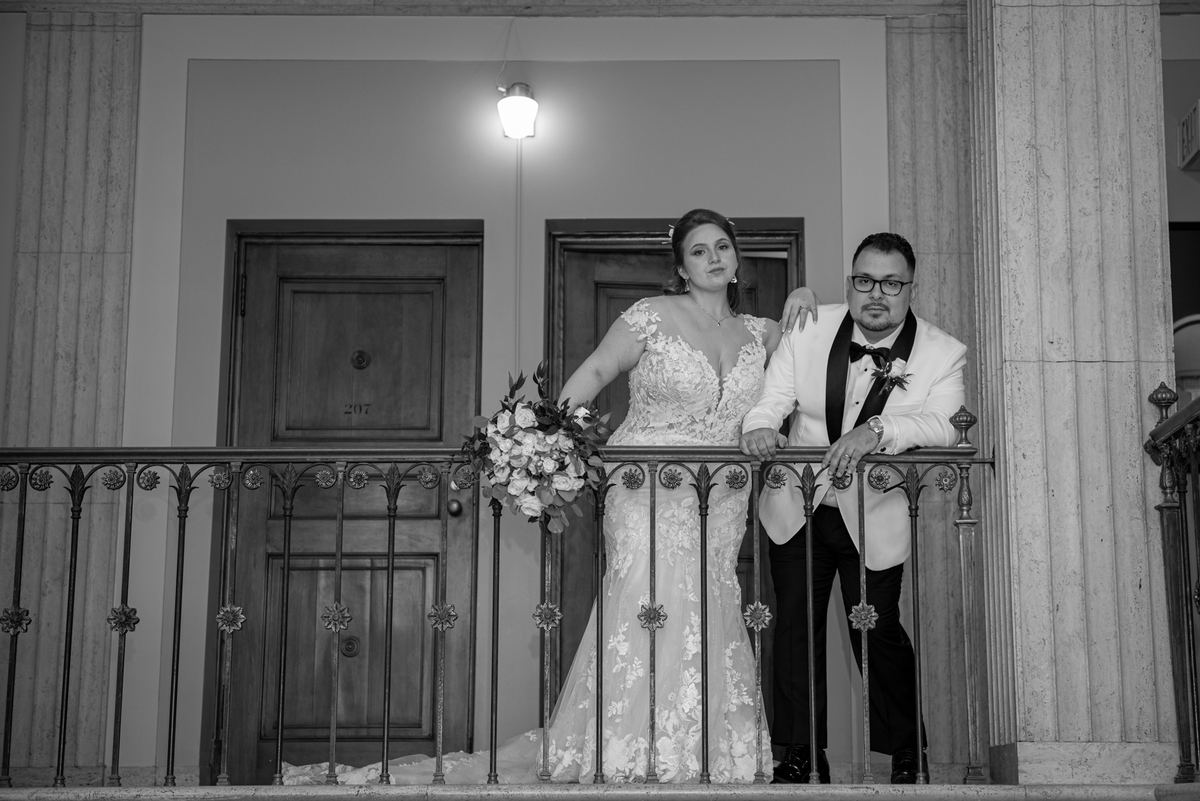 A bride and groom stand on an ornate balcony, dressed formally against a backdrop of classical doors and columns.