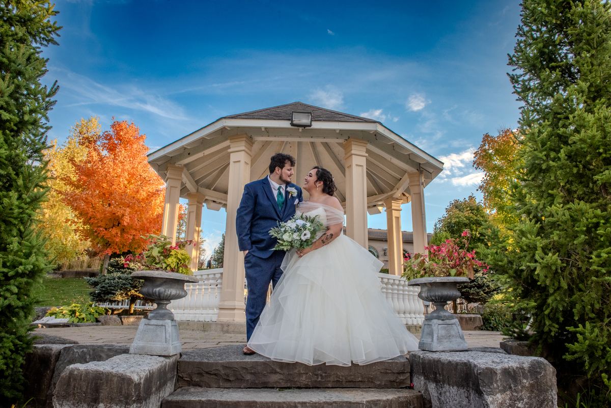 A smiling bride and groom share an intimate glance on stone steps, surrounded by autumn foliage under a bright blue sky.