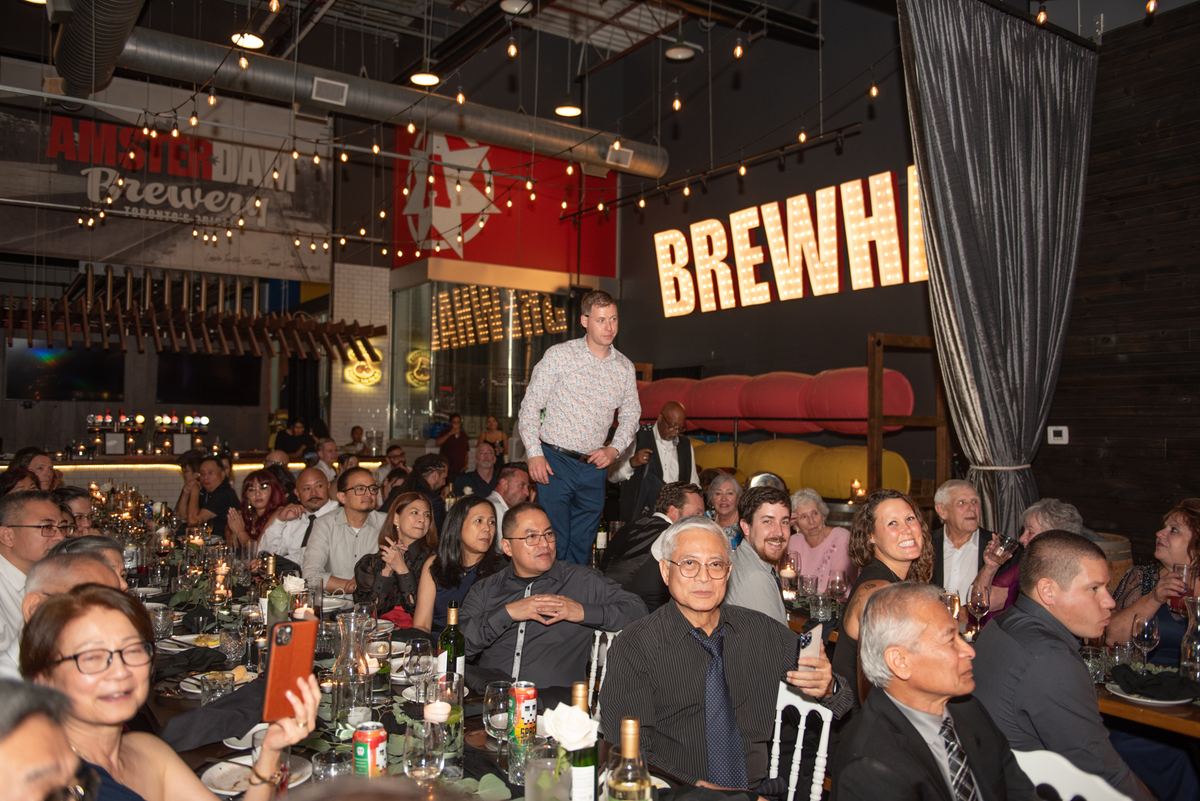 People gather at long tables in an industrial-chic brewery, watching a speaker under string lights and the BREWHA sign.