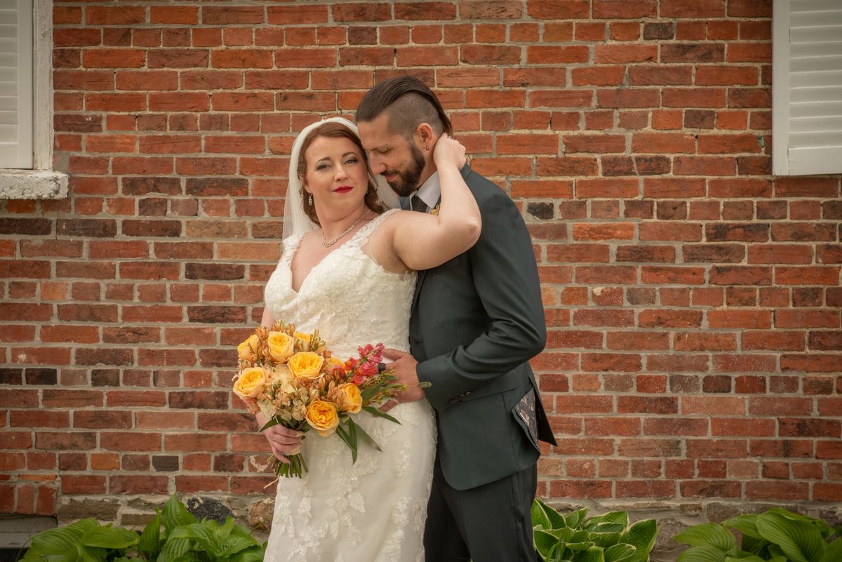 A bride with a yellow and orange bouquet is tenderly embraced by her groom against a rustic brick wall.