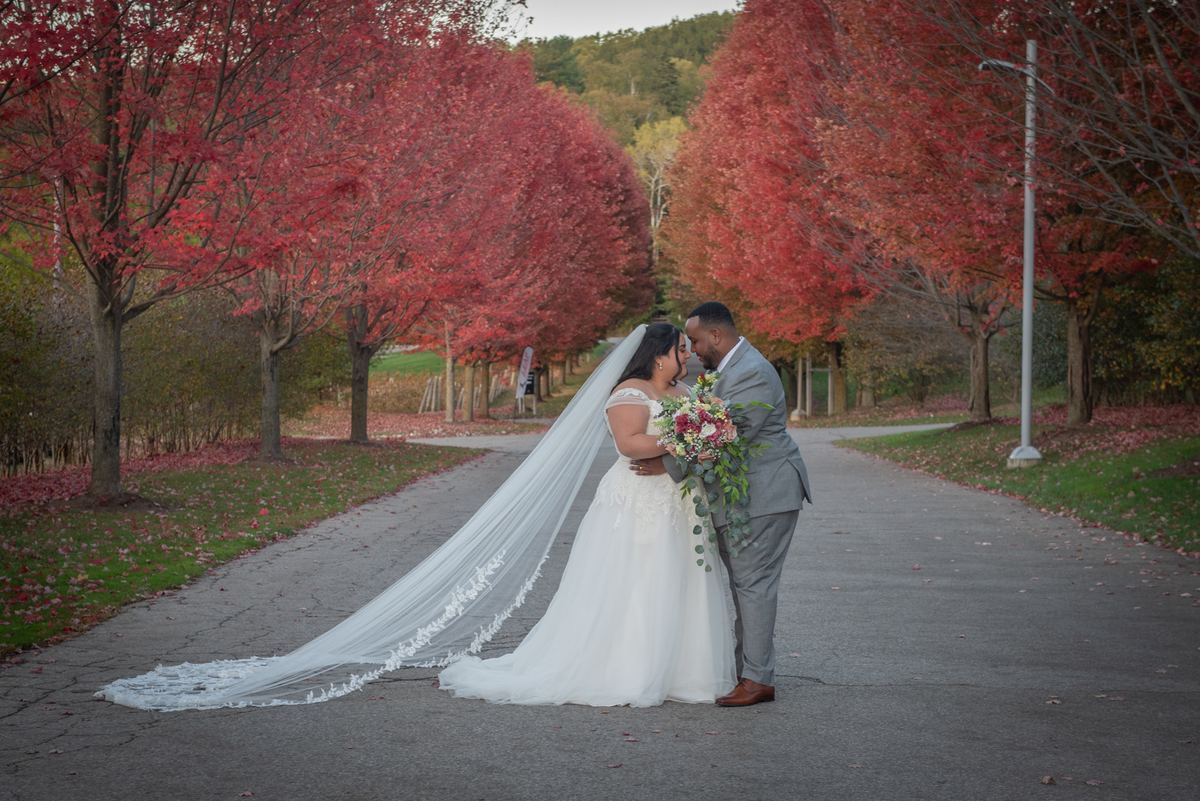 A newlywed couple stands close, foreheads touching under autumn's fiery canopy, glowing in late afternoon light.