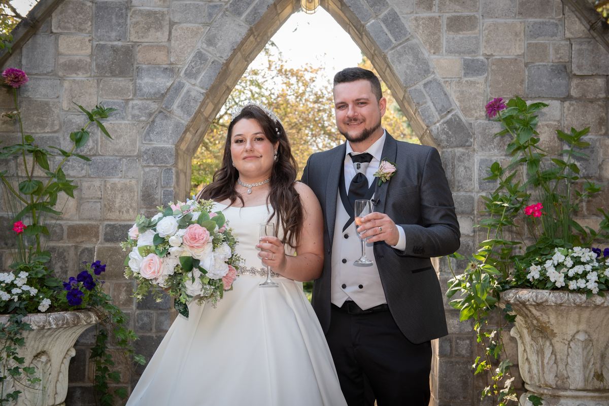 A bride and groom smile under a stone archway, holding champagne flutes surrounded by greenery and flowers.